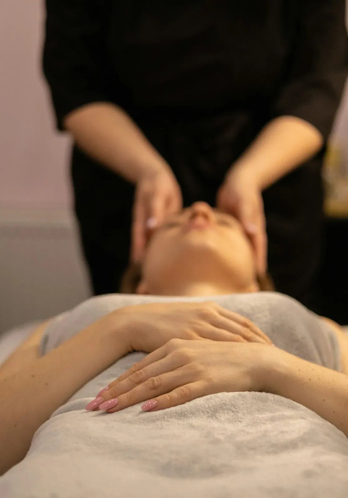 Mulher deitada em uma maca de olhos fechados recebendo massagem facial das mãos de uma terapeuta